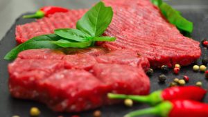 Carne equina fresca su tagliere con foglie di basilico, peperoncini rossi e pepe misto, pronta per la preparazione